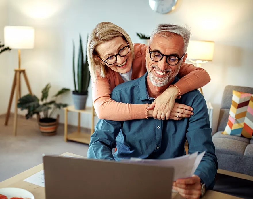 Retired couple hugging and laughing while browsing on their laptop